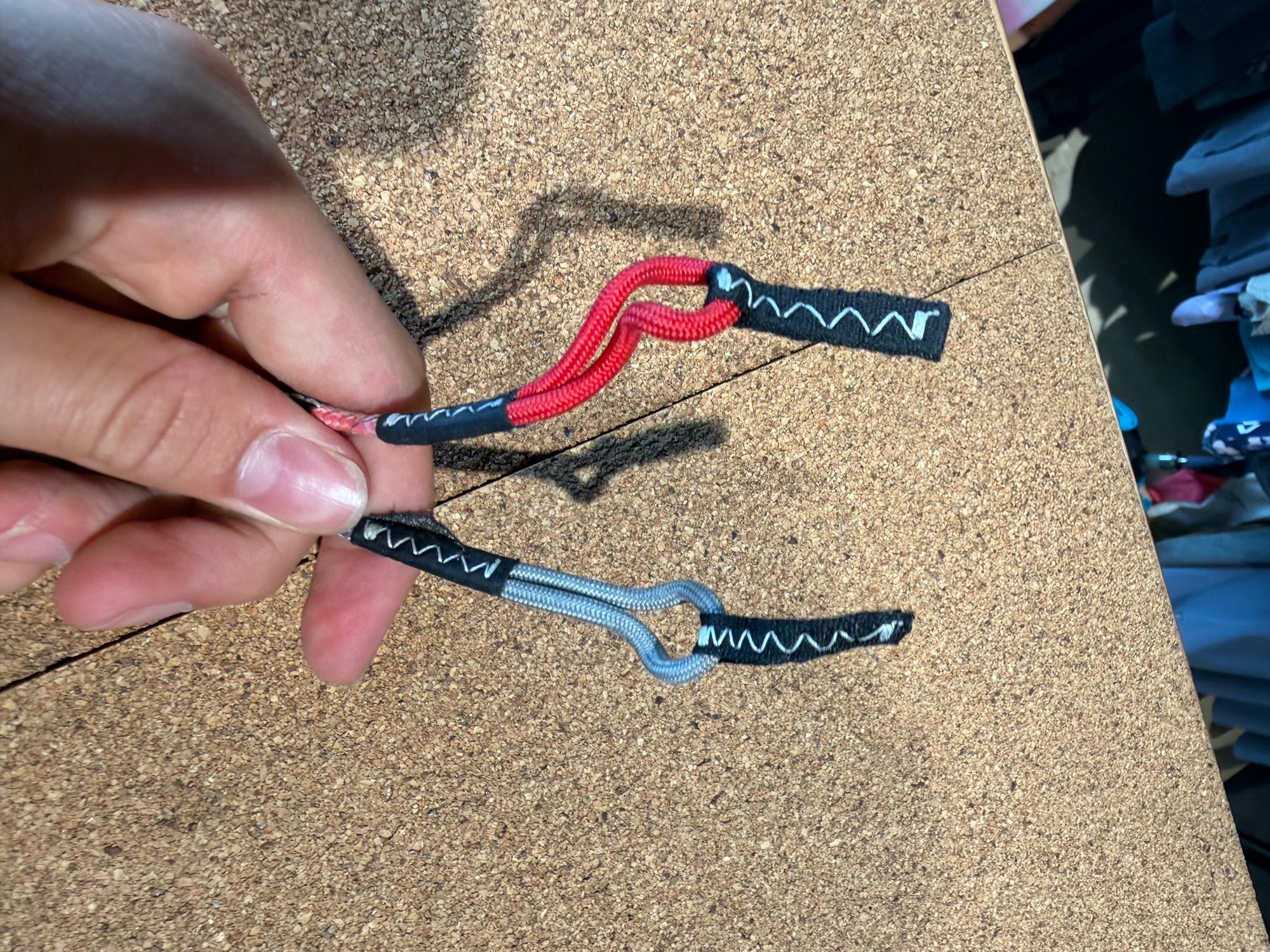 Hand holding red and grey Duotone Click Bar pigtail connectors with black stitched reinforced loops on a cork background.