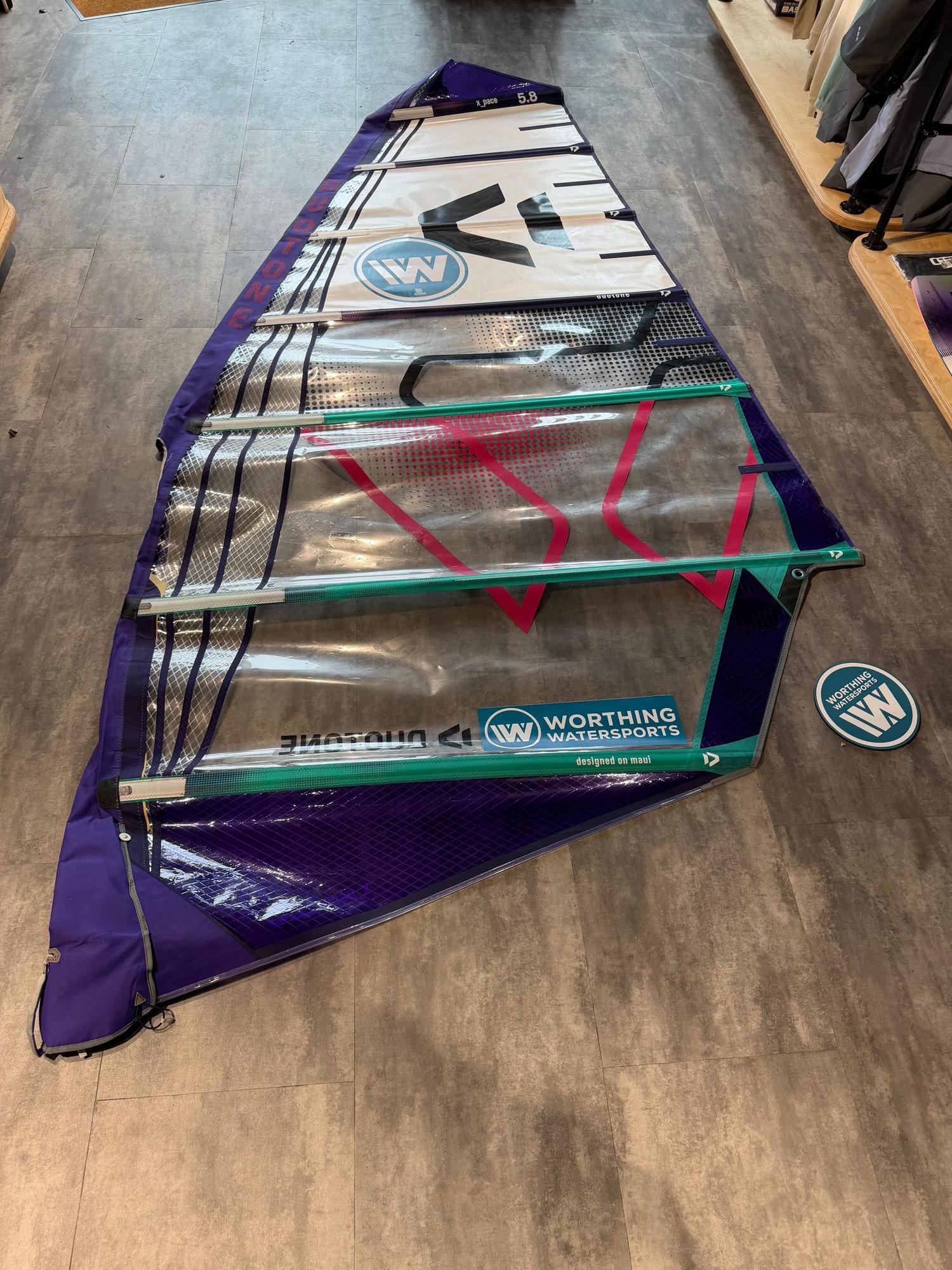 Used Duotone X_Pace 5.8 freerace windsurf sail in purple and white with teal detailing, laid flat on a shop floor.
