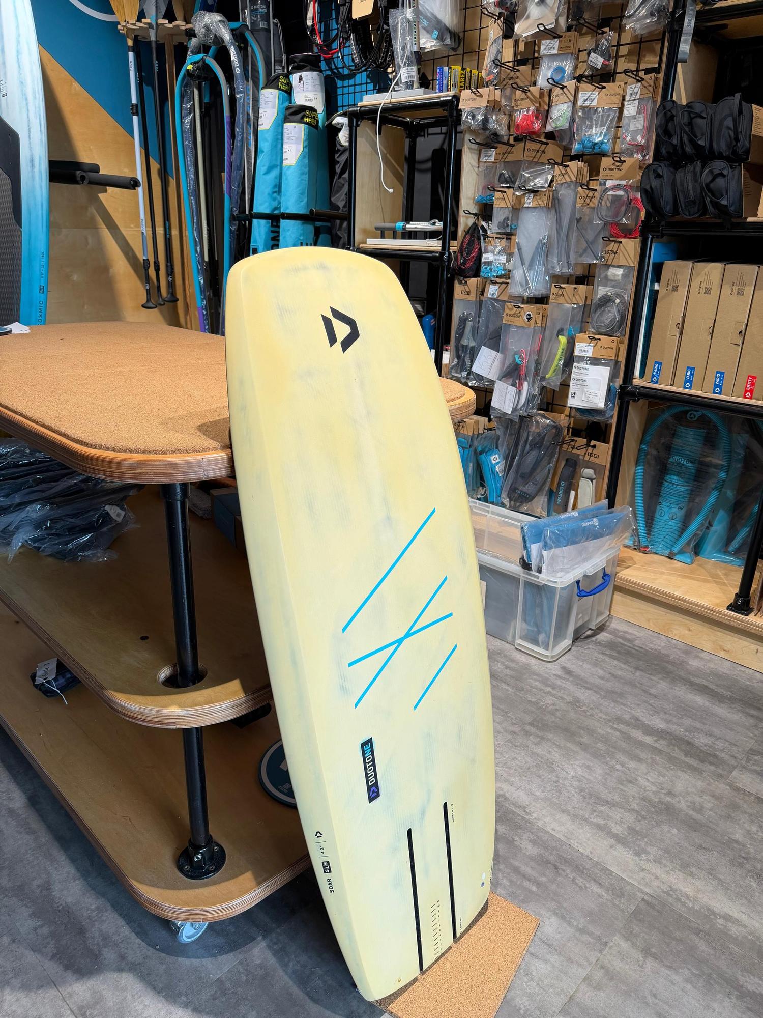 Used Duotone Soar SLS 4'2'' kite foilboard in yellow carbon with blue graphics, standing upright in a retail store.
