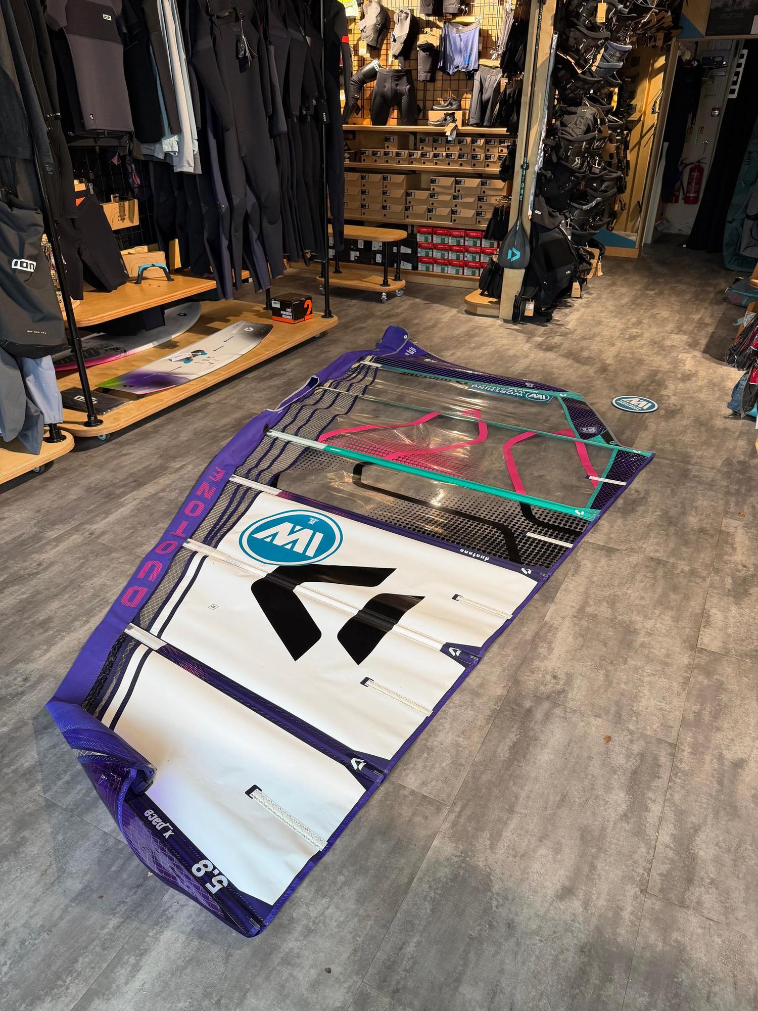 Used Duotone X_Pace 5.8 freerace windsurf sail in purple, white, and clear monofilm, laid flat on a shop floor.