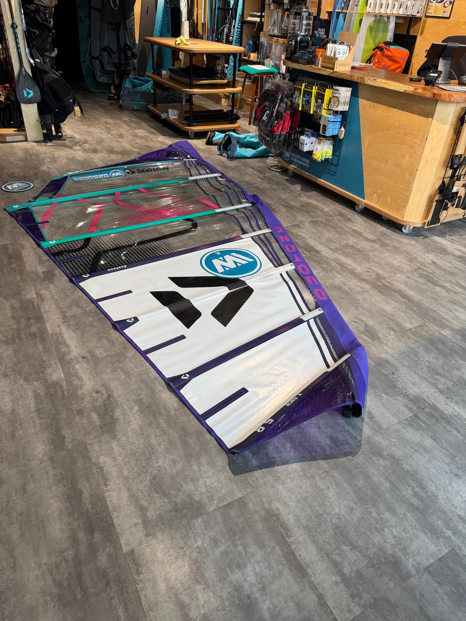 Used Duotone X_Pace 5.8 freerace windsurf sail in purple, white, and teal, laid flat on a shop floor.