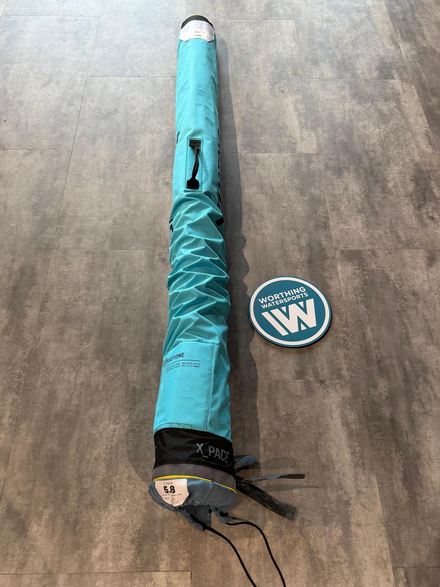 Used Duotone X_Pace 5.8 freeride windsurf sail rolled in a teal storage bag on a grey floor.
