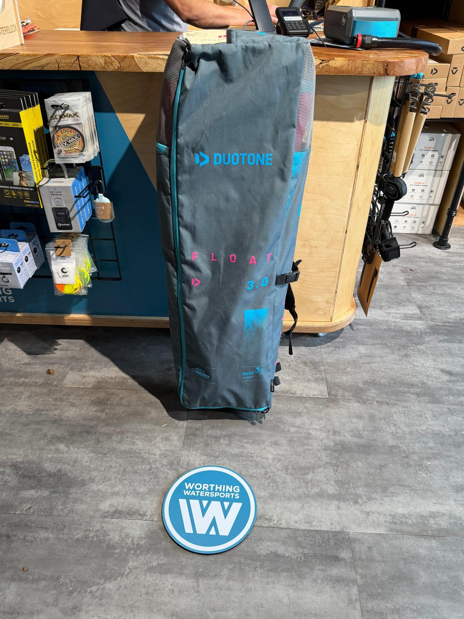 Grey Duotone Float 3m wave wing in its branded storage bag with blue and pink detailing, standing on a shop floor.