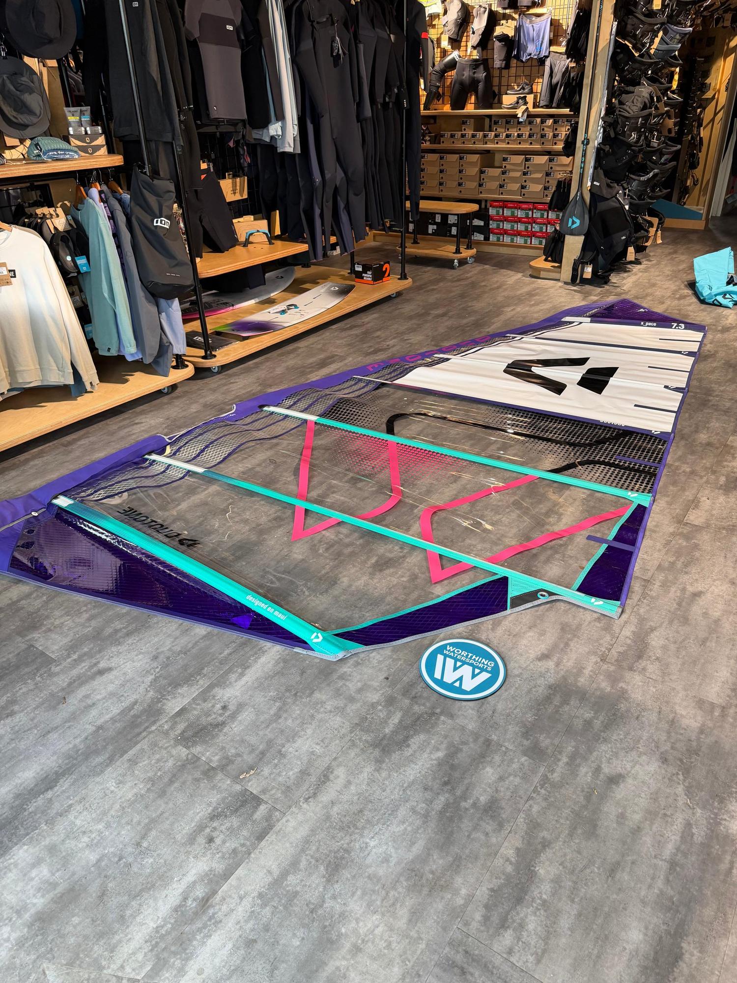 Used Duotone X_Pace 7.3m freerace windsurf sail in purple, white, and teal, laid flat on a shop floor.
