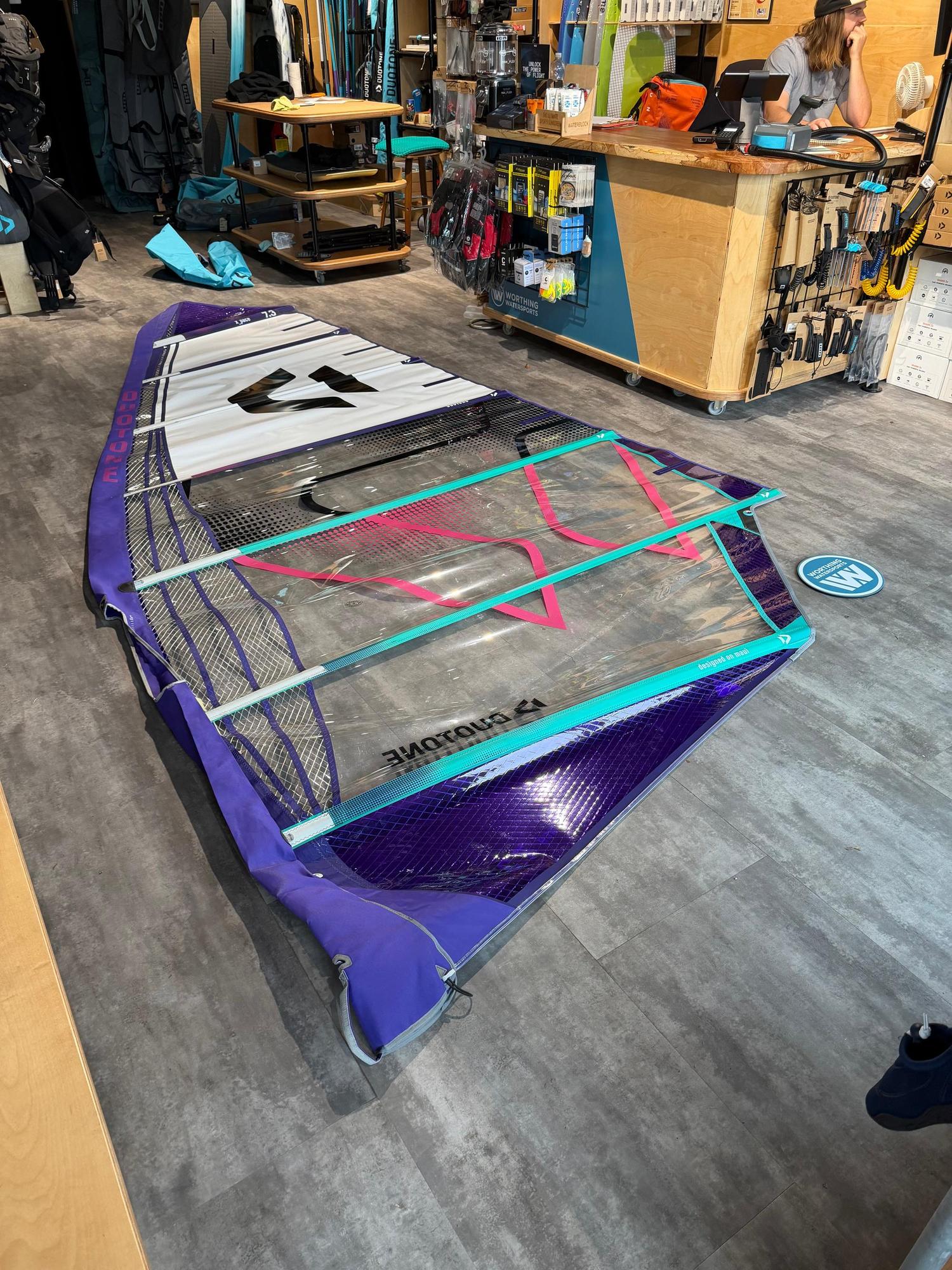 Used Duotone X_Pace 7.3m freerace windsurf sail in purple, white, and clear monofilm with pink detailing laid on a shop floor.