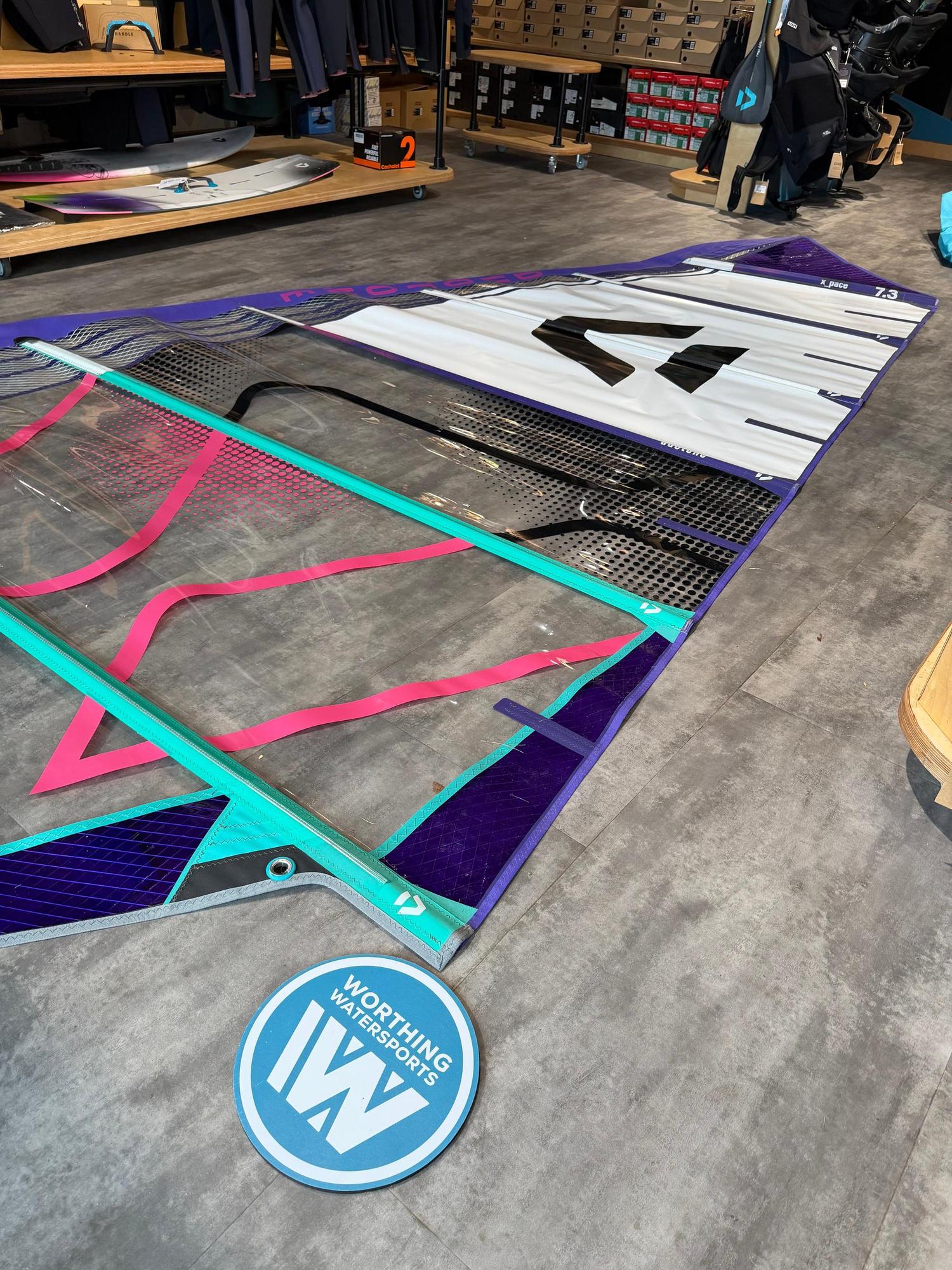 Used Duotone X_Pace 7.3m freerace windsurf sail in purple, white, and teal, laid flat on a shop floor.